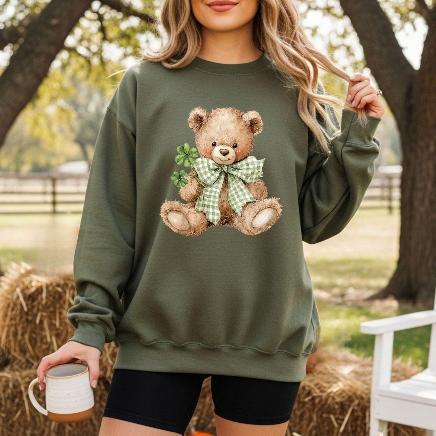 Woman in a green sweatshirt with a watercolor teddy bear holding four-leaf clovers and wearing a shamrock bow.