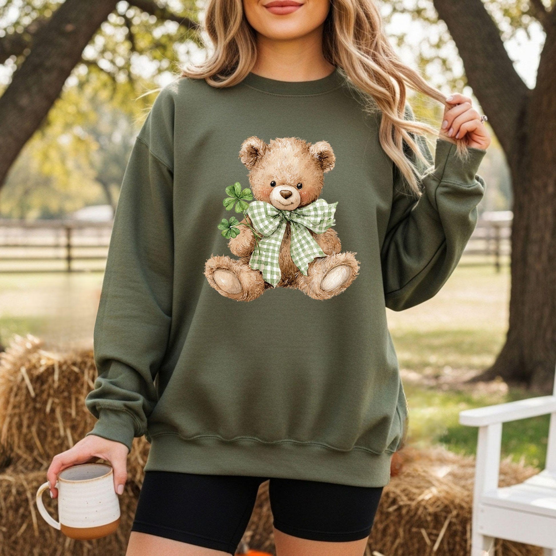 Woman in a green sweatshirt with a watercolor teddy bear holding four-leaf clovers and wearing a shamrock bow.