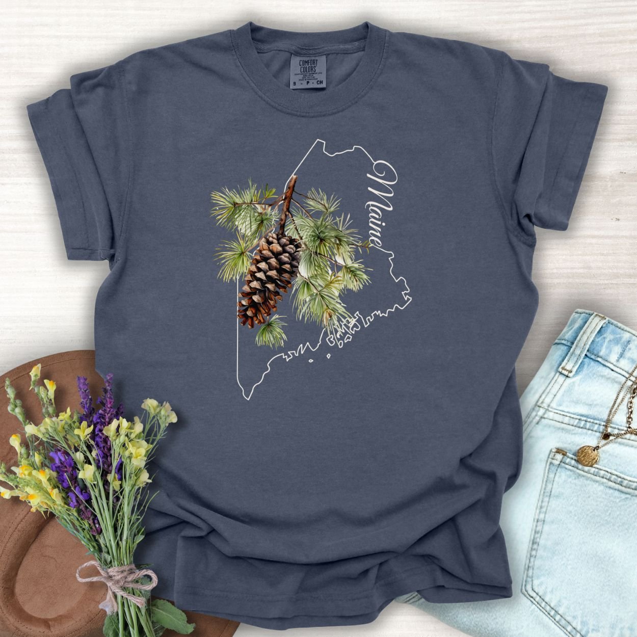 Denim blue garment-dyed t-shirt with a white Maine outline and a pine cone and branch graphic.