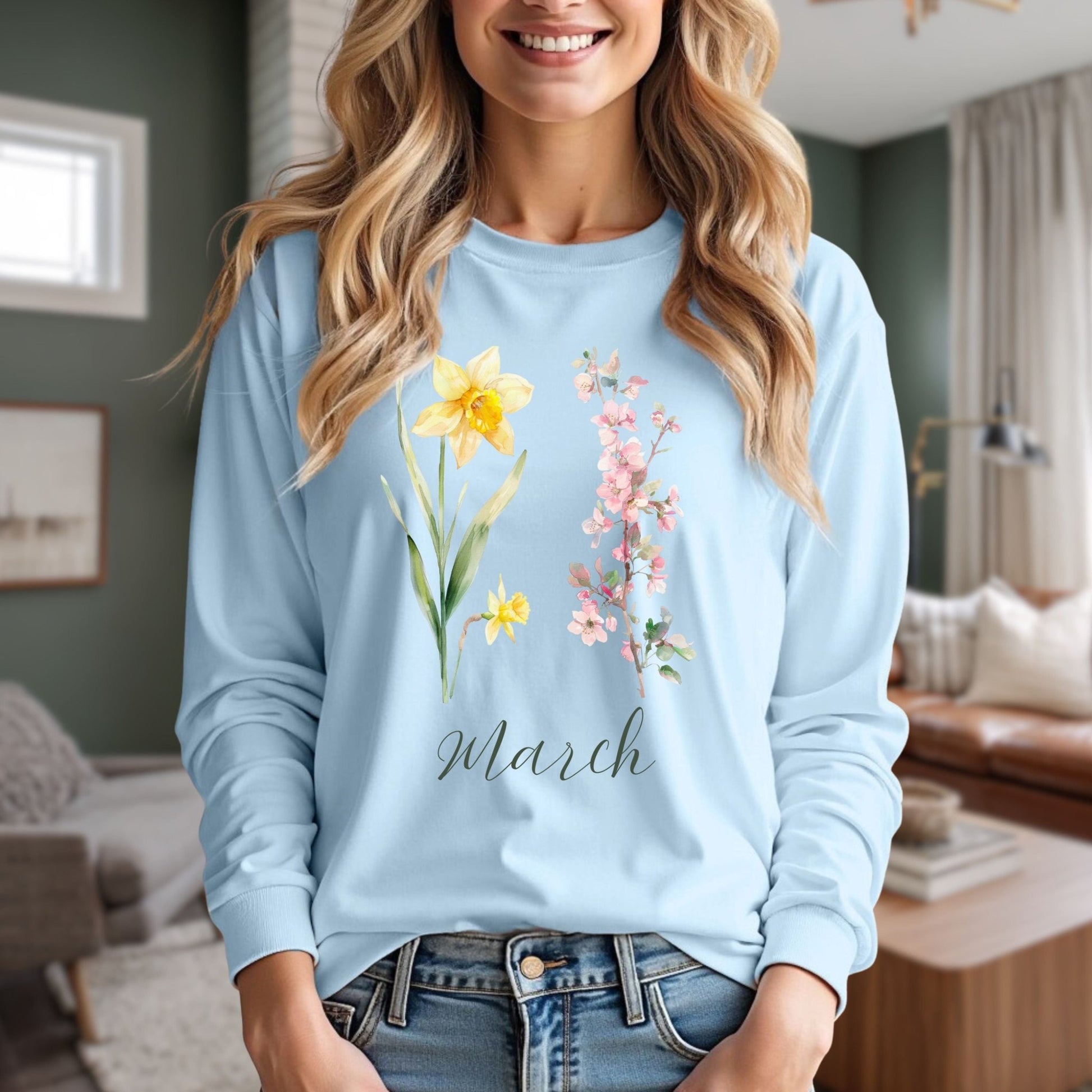 Smiling woman in a light blue long-sleeve tee with hand-drawn daffodils, pink cherry blossoms, and "March" script.