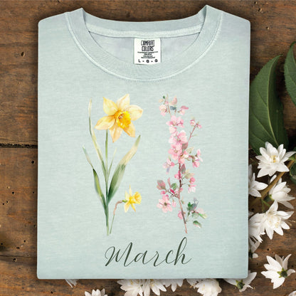 Light blue garment-dyed cotton t-shirt with watercolor daffodils, cherry blossoms, and "March" script.