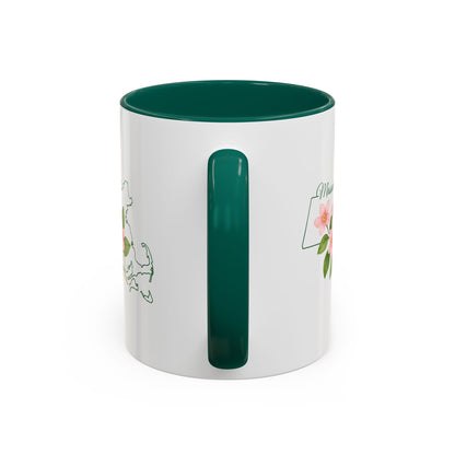 White ceramic mug with a green handle and interior, featuring a Massachusetts state outline and pink Mayflower blossoms.