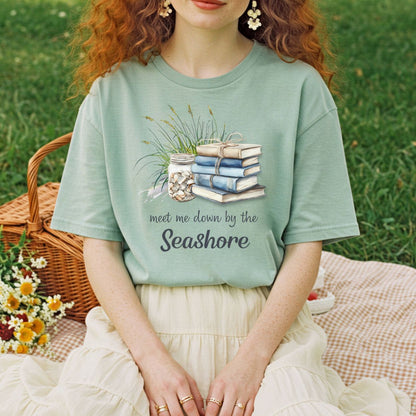 Garment-dyed sage green t-shirt with "Meet Me Down By the Seashore" text, books, shells, and dune grass graphic.