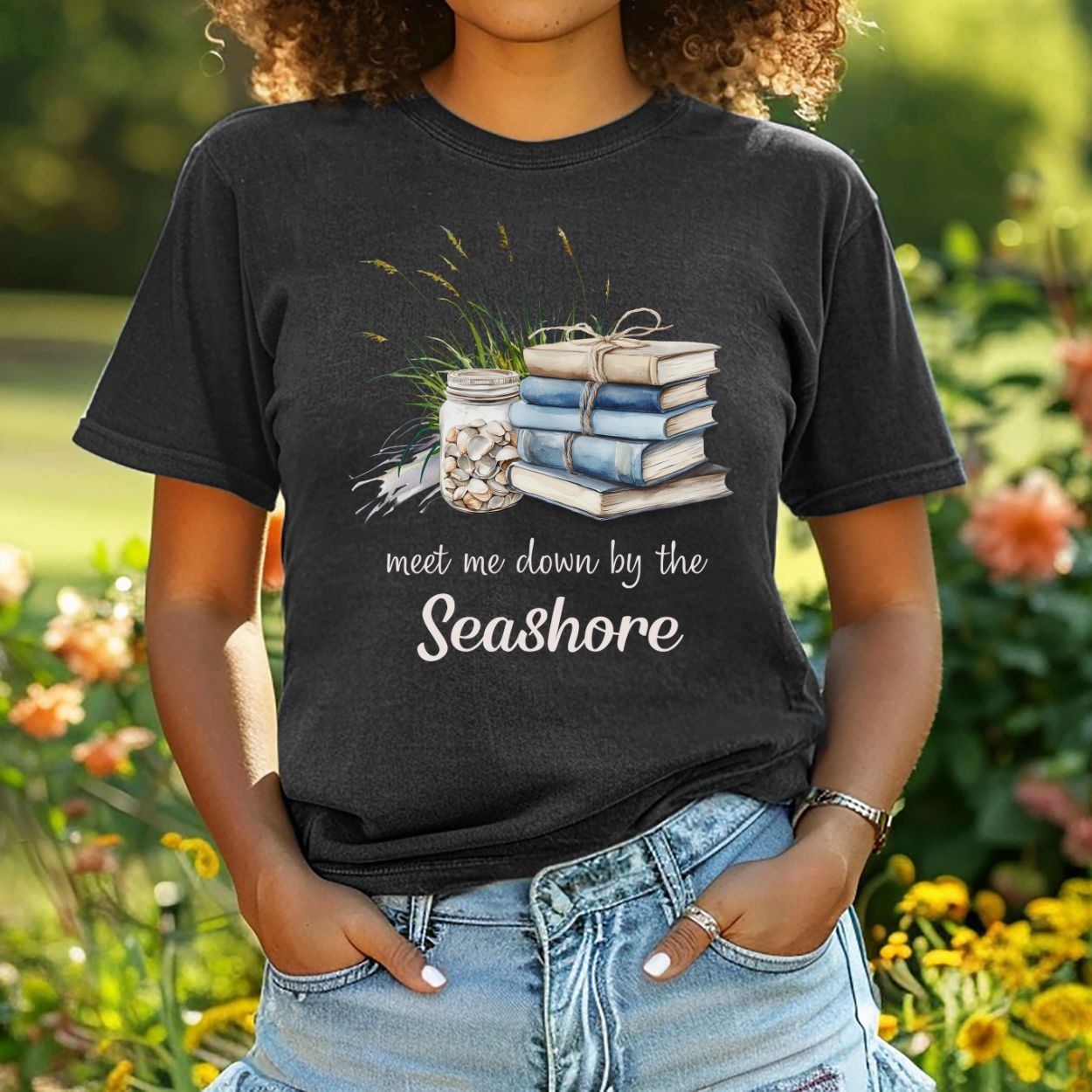Black garment-dyed t-shirt with "meet me down by the Seashore" text, stacked books, shells, and dune grass design.