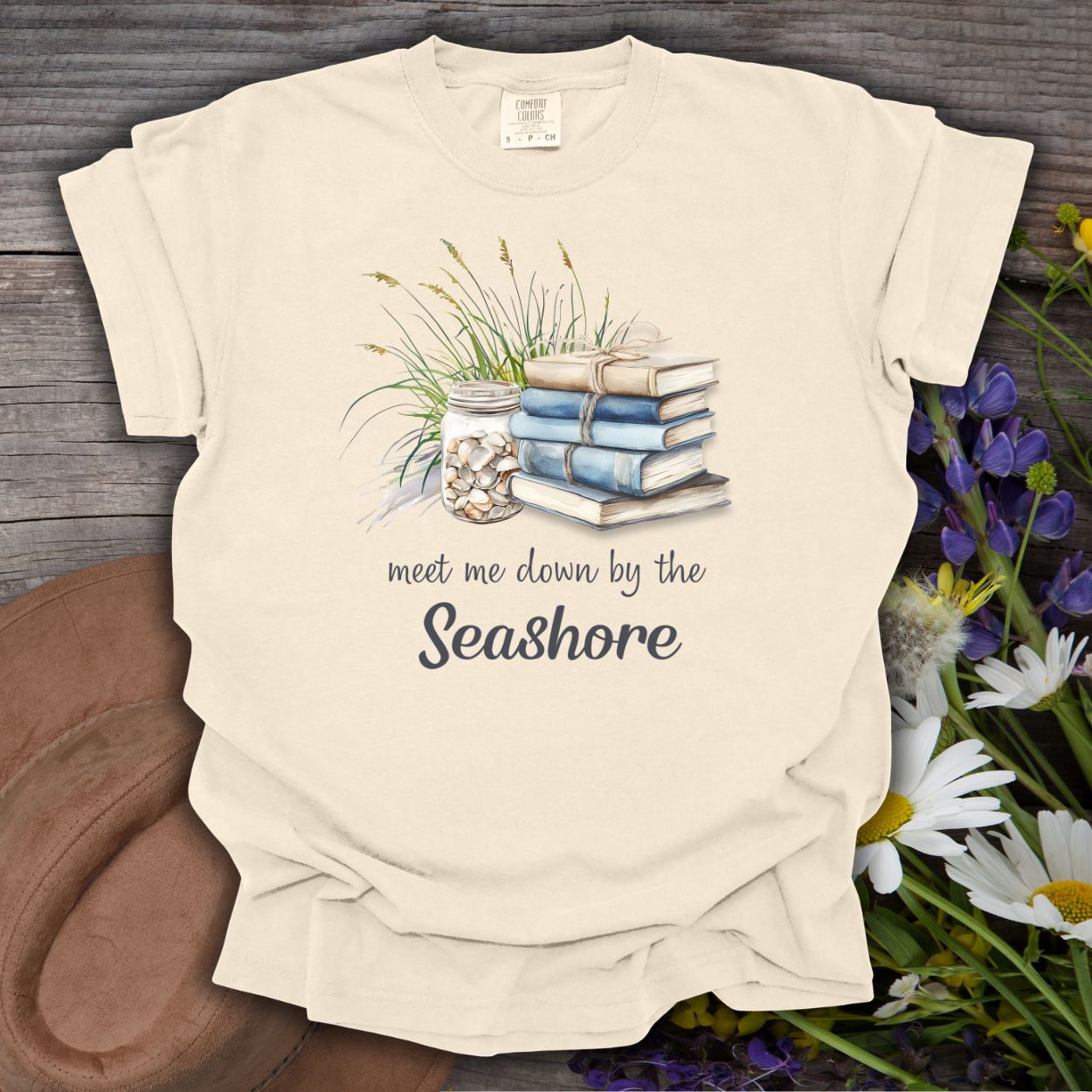 Ivory garment-dyed cotton t-shirt with "Meet Me Down By the Seashore" text, books, shells, and grass graphic.