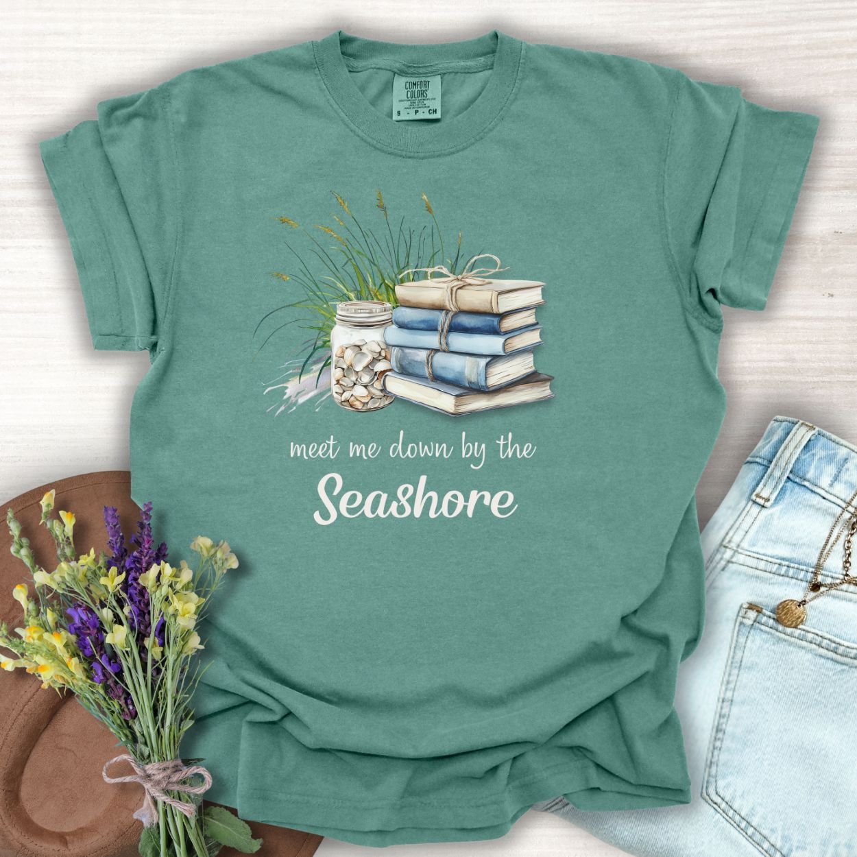 Light green S t-shirt with "meet me down by the Seashore" text, stacked books, shells, and dune grass.