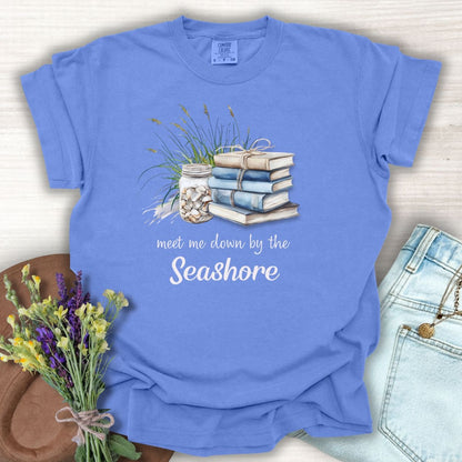 Periwinkle garment-dyed t-shirt with "meet me down by the Seashore" text, stacked books, shells, and dune grass.