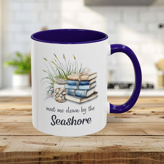 White ceramic 11oz mug with blue interior/handle, featuring watercolor stacked books, beach grasses, shells, and "meet me down by the Seashore" text.