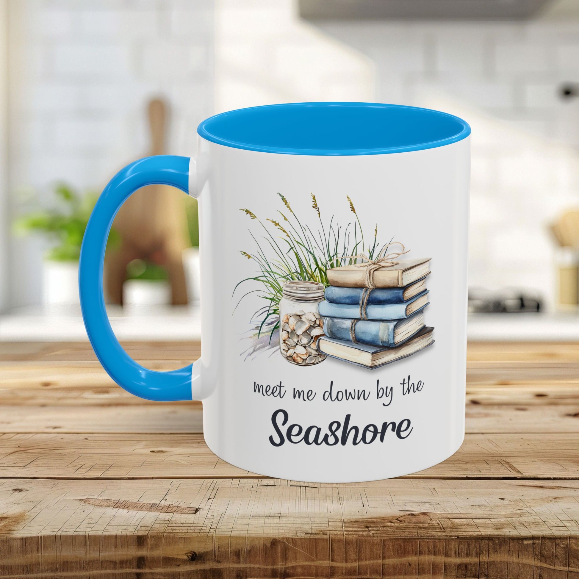 White ceramic mug with blue handle and interior, featuring watercolor stacked books, grasses, and shells, with "meet me down by the Seashore" text.