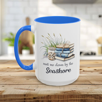 White ceramic mug with blue handle and interior, featuring watercolor art of stacked books, beach grasses, and shells.