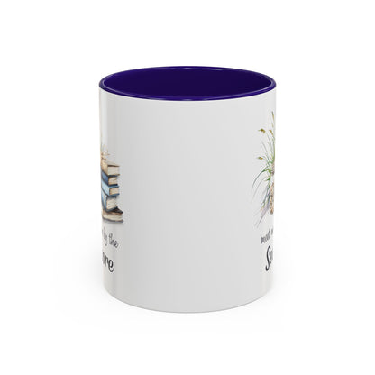 White ceramic mug with a purple interior and handle, featuring watercolor art of stacked books, beach grasses, and shells.