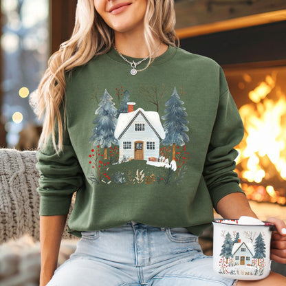Cozy green crewneck sweatshirt with a snowy white cabin and pine trees, held by a blonde woman.