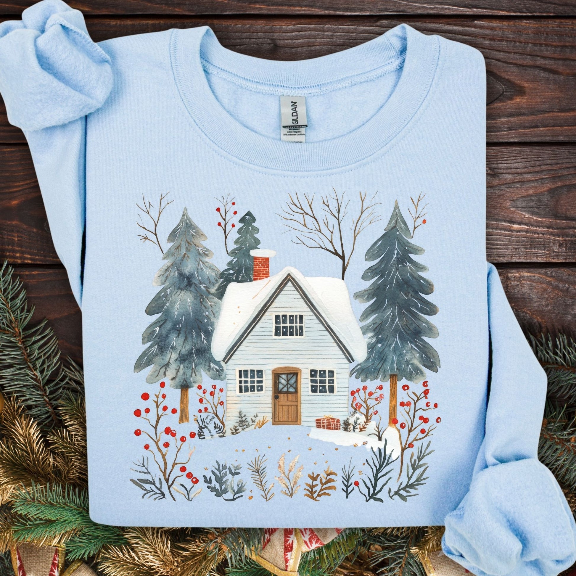 Light blue crewneck sweatshirt with a snowy cabin, pine trees, and red berries in folk-art style.