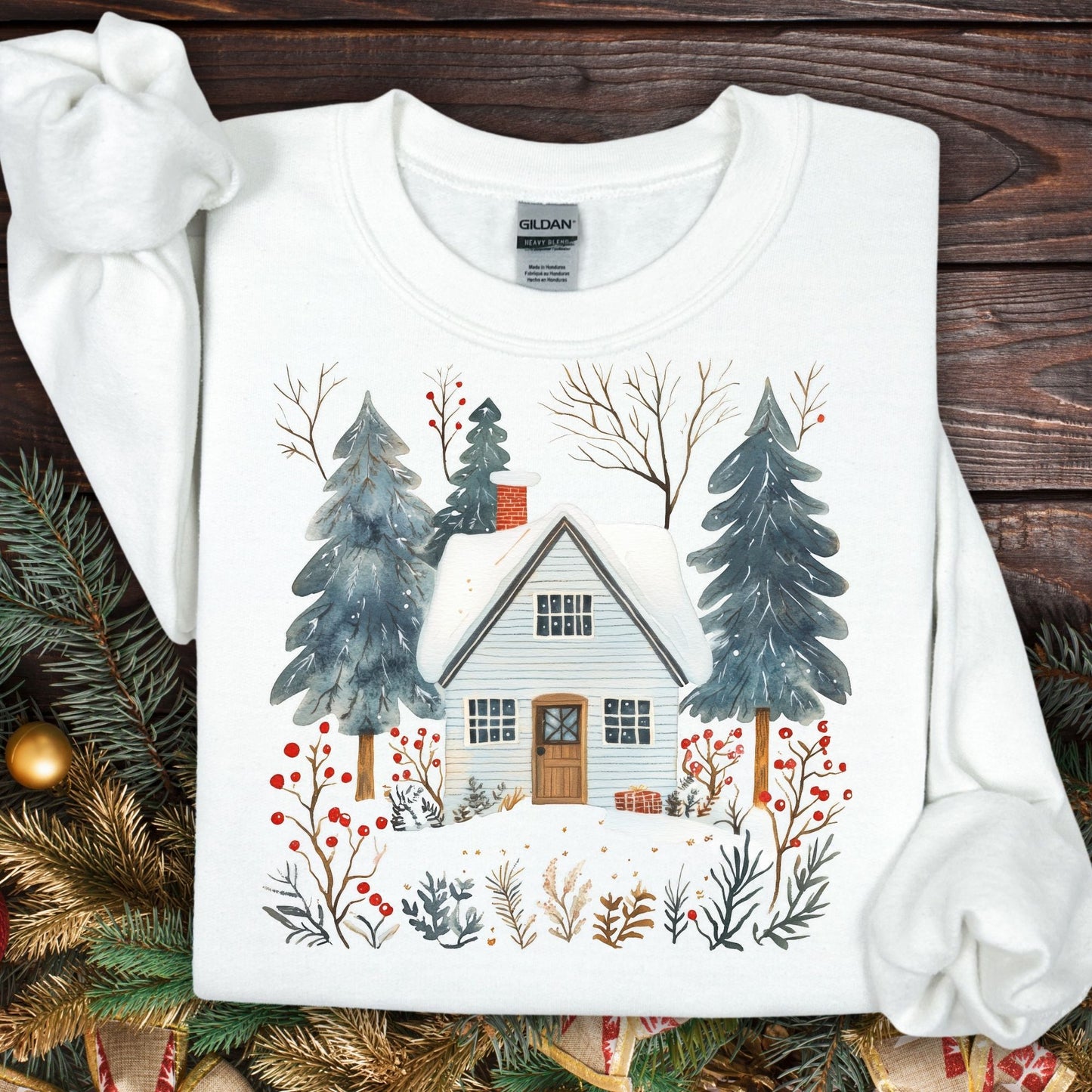 White Nordic Winter Cabin Sweatshirt with snowy cabin, pine trees, and red berries, on a wooden background.
