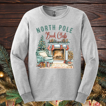 Ash Gildan long sleeve tee with "North Pole Book Club" text and a cozy fireplace, armchair, and Christmas tree design.