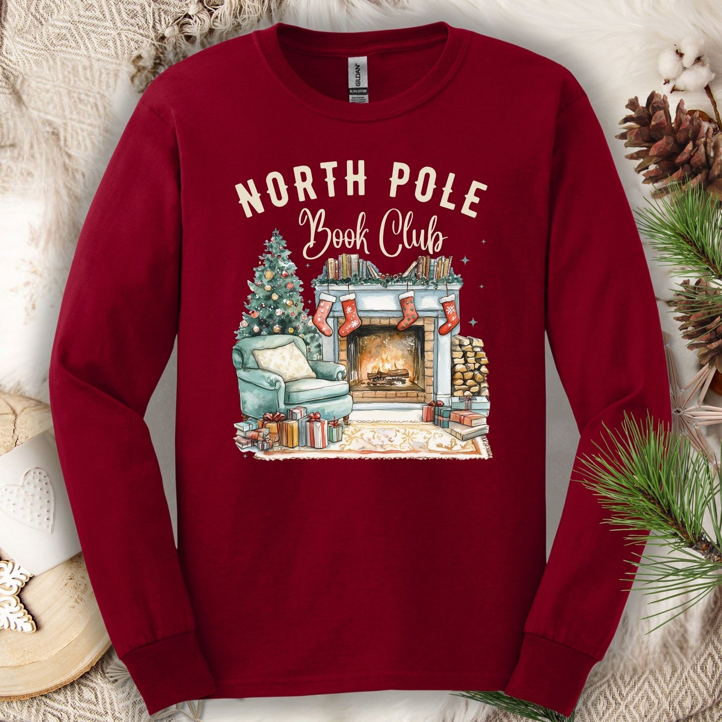 Cardinal Red Gildan long sleeve tee with "North Pole Book Club" text and a cozy fireplace, armchair, and Christmas tree graphic.