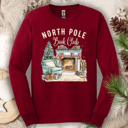 Cardinal Red Gildan long sleeve tee with "North Pole Book Club" text and a cozy fireplace, armchair, and Christmas tree graphic.