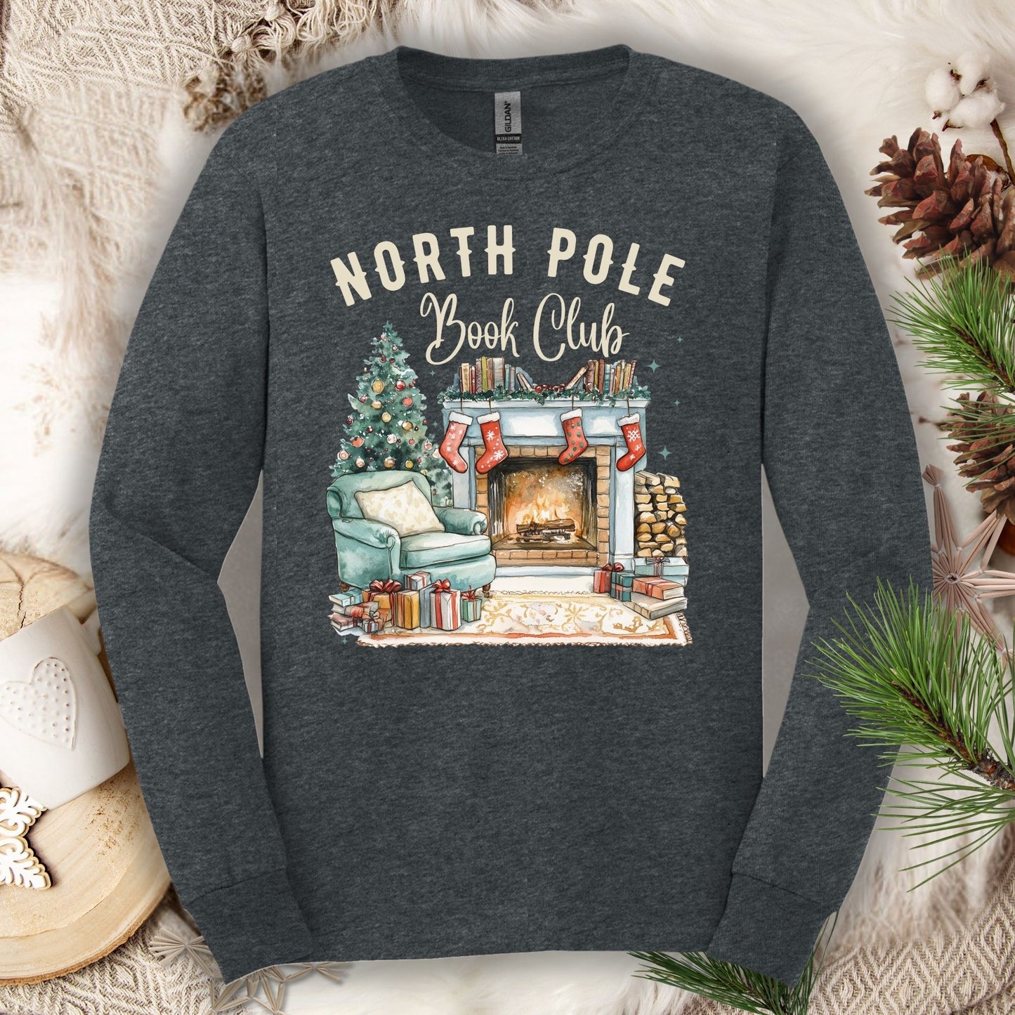 Dark Heather Gildan long sleeve tee with "North Pole Book Club" text above a cozy fireplace, armchair, and Christmas tree.