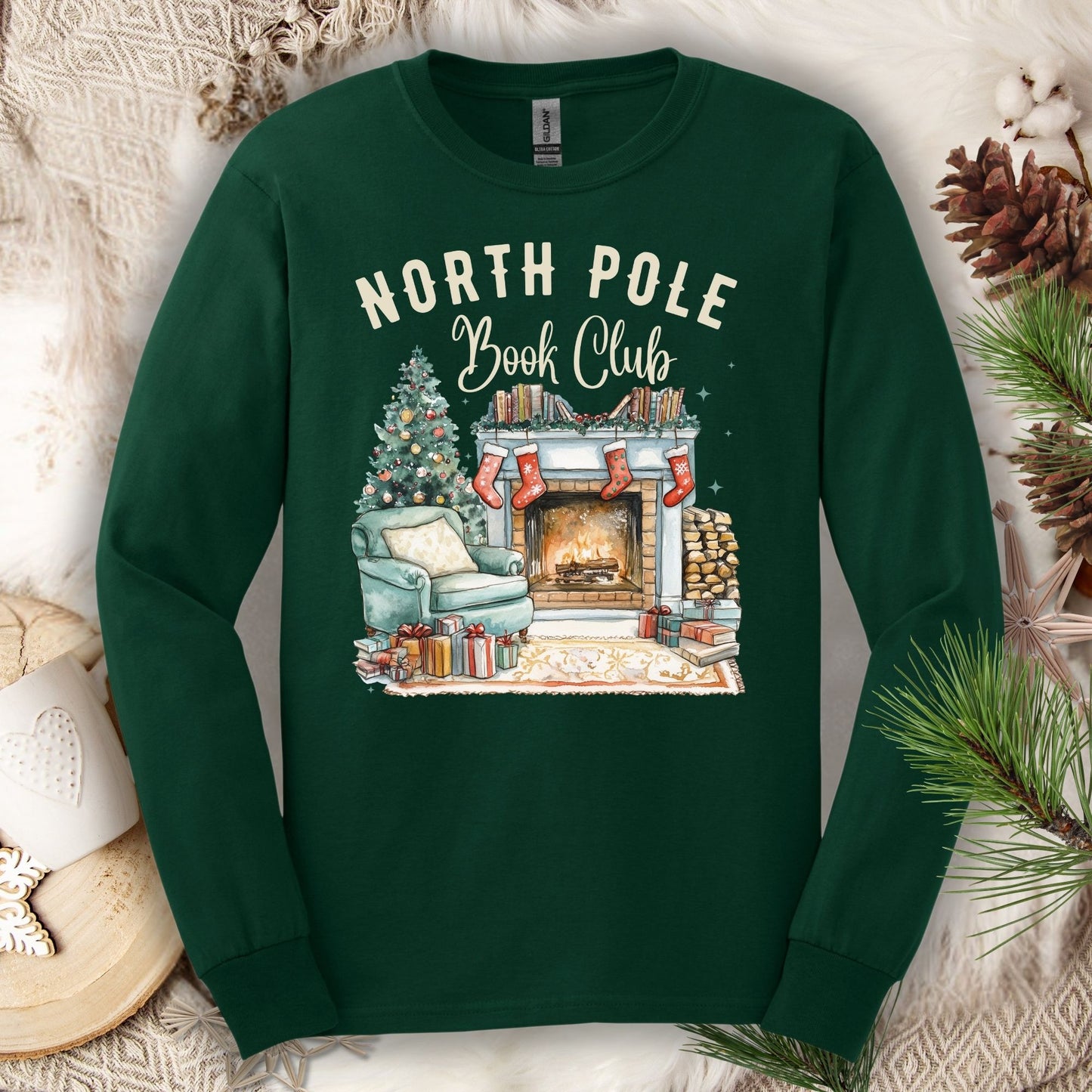 Forest Green Gildan long sleeve tee with "North Pole Book Club" text above a cozy fireplace, armchair, and Christmas tree scene.