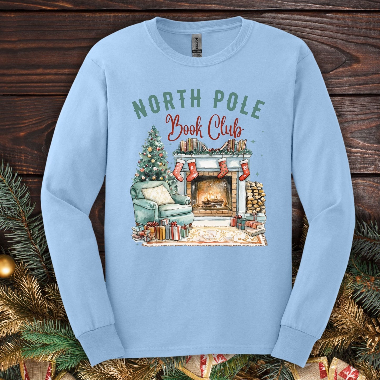 Light blue Gildan long sleeve tee with "North Pole Book Club" and a cozy Christmas fireplace scene.