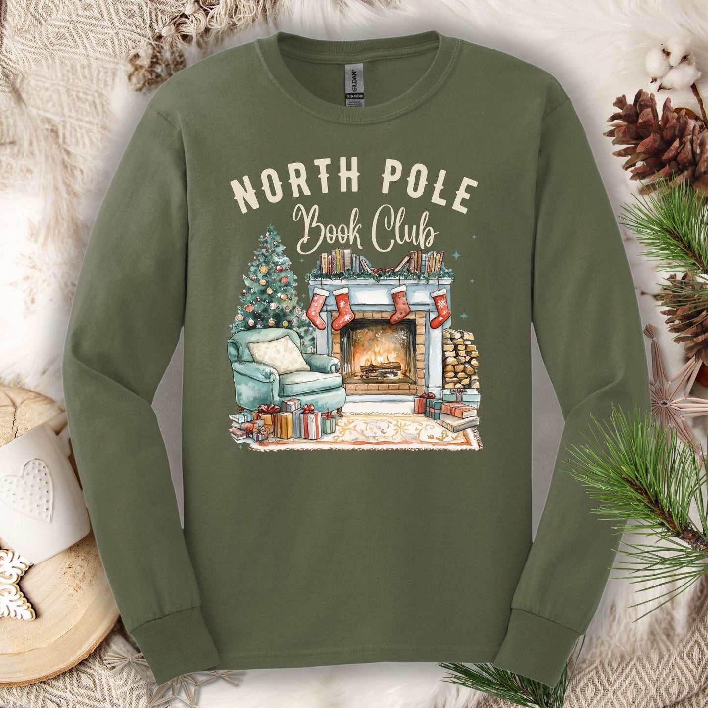 Military Green Gildan long sleeve tee with "North Pole Book Club" text and a cozy Christmas fireplace scene.