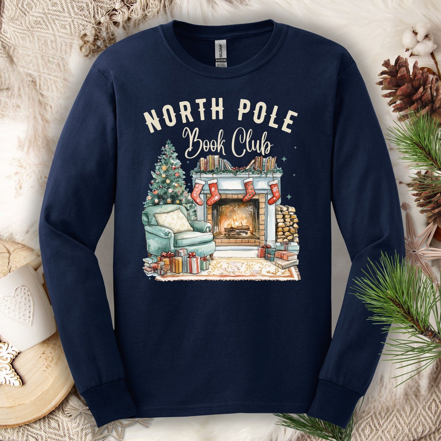 Navy Gildan long sleeve tee with "North Pole Book Club" text and a cozy fireplace, armchair, and Christmas tree design.