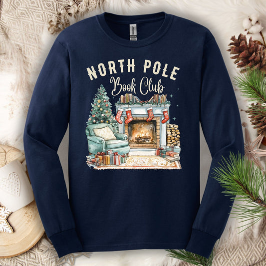 Navy Gildan long sleeve tee with "North Pole Book Club" text and a cozy fireplace, armchair, and Christmas tree design.