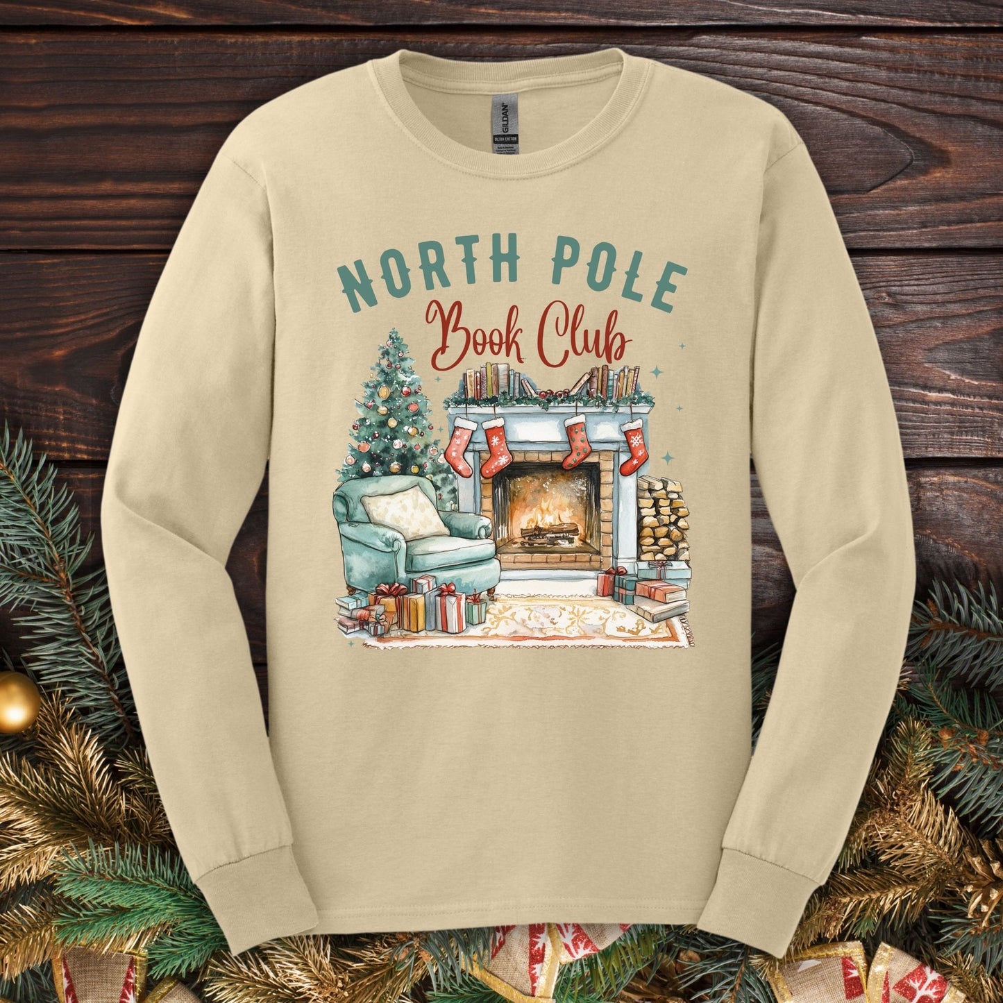 Sand Gildan long sleeve tee with a "North Pole Book Club" fireplace, armchair, and Christmas tree graphic.