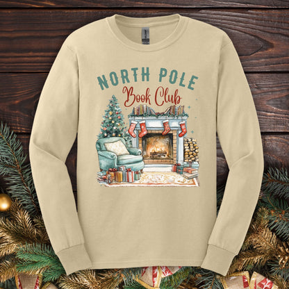 Sand Gildan long sleeve tee with a "North Pole Book Club" fireplace, armchair, and Christmas tree graphic.