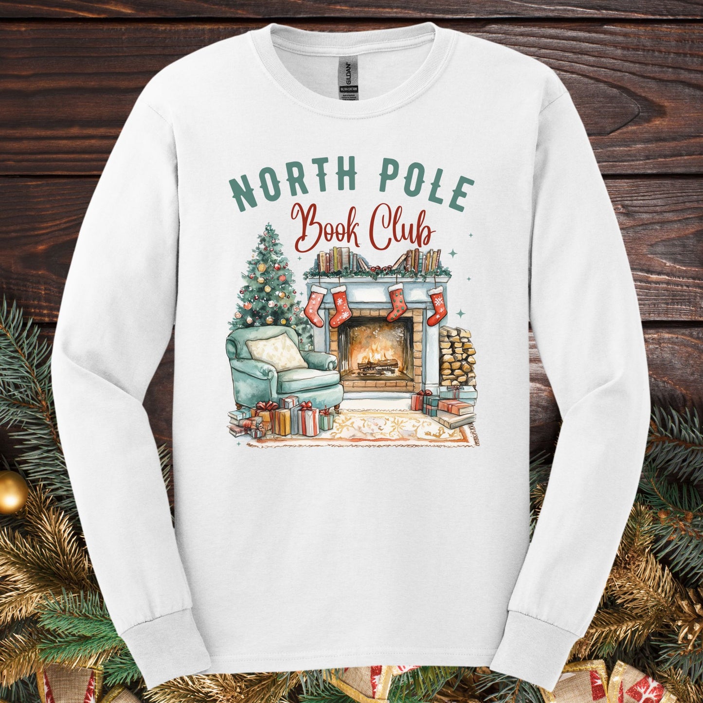 White Gildan long sleeve tee with "North Pole Book Club" text and a cozy fireplace, armchair, and Christmas tree design.