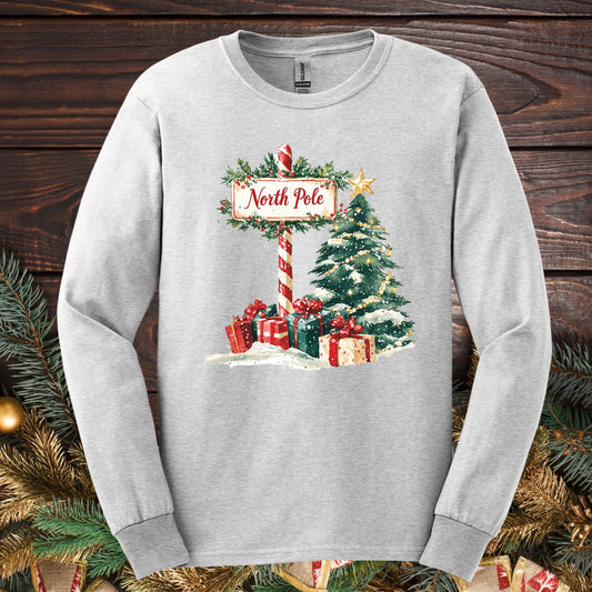 Ash long sleeve tee with a watercolor North Pole scene, gifts, and a glowing Christmas tree.