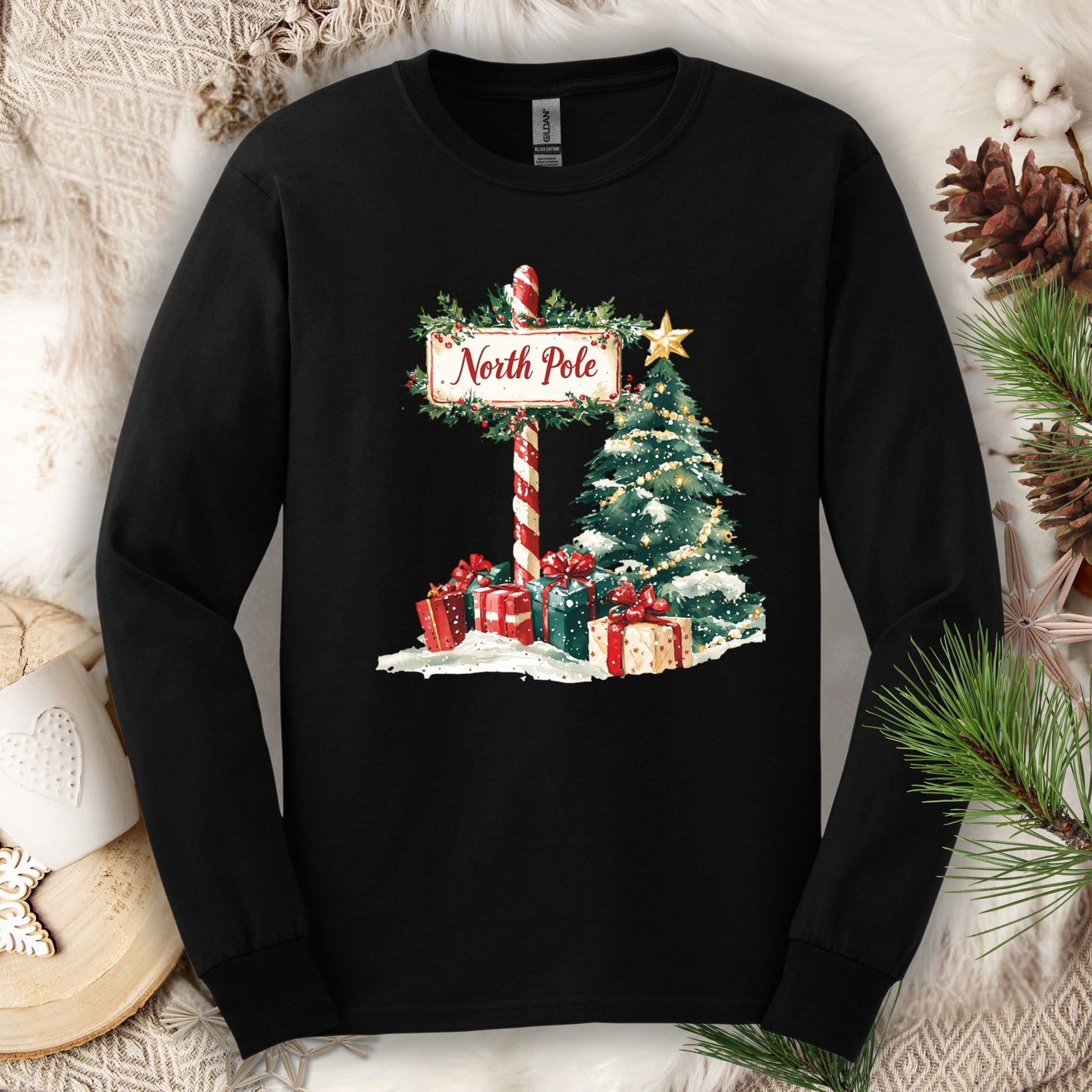Black Gildan long sleeve tee with a watercolor North Pole scene: snow, gifts, and a glowing Christmas tree.