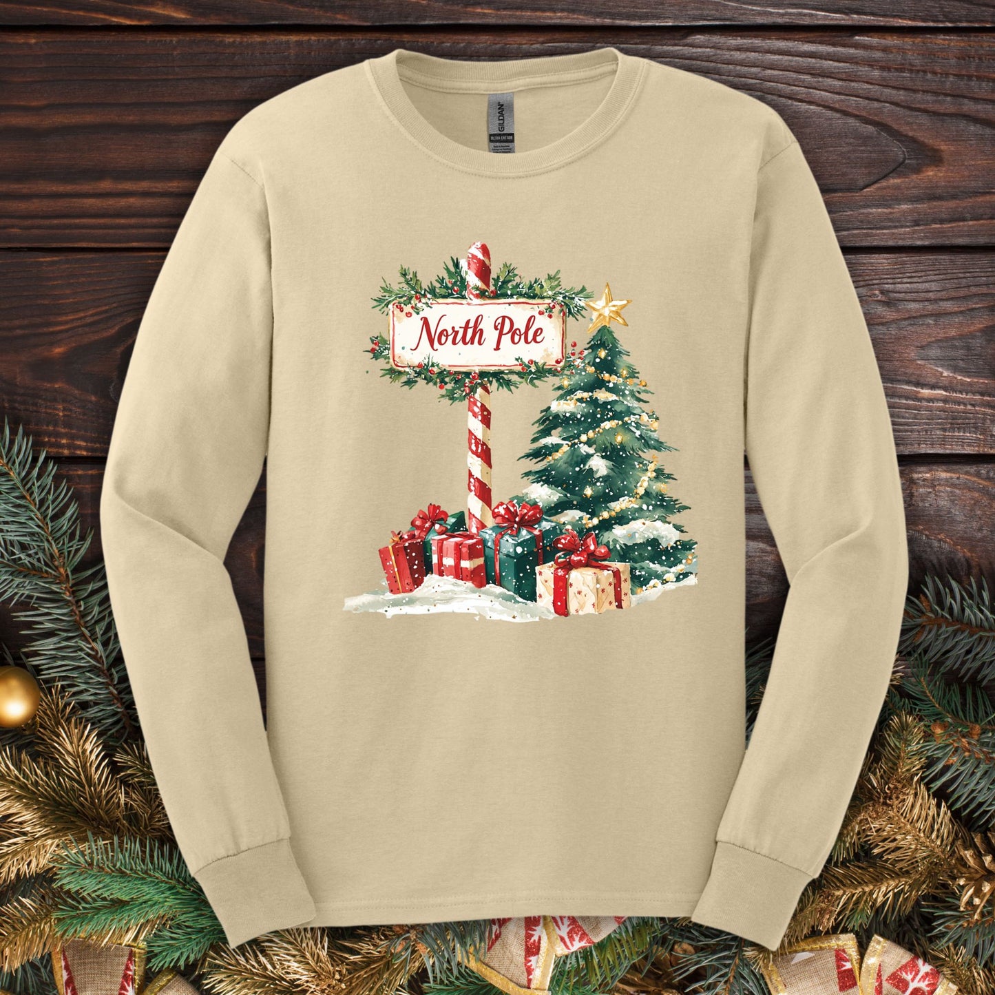 Sand Gildan long sleeve tee with a watercolor North Pole sign, snowy tree, and wrapped gifts.