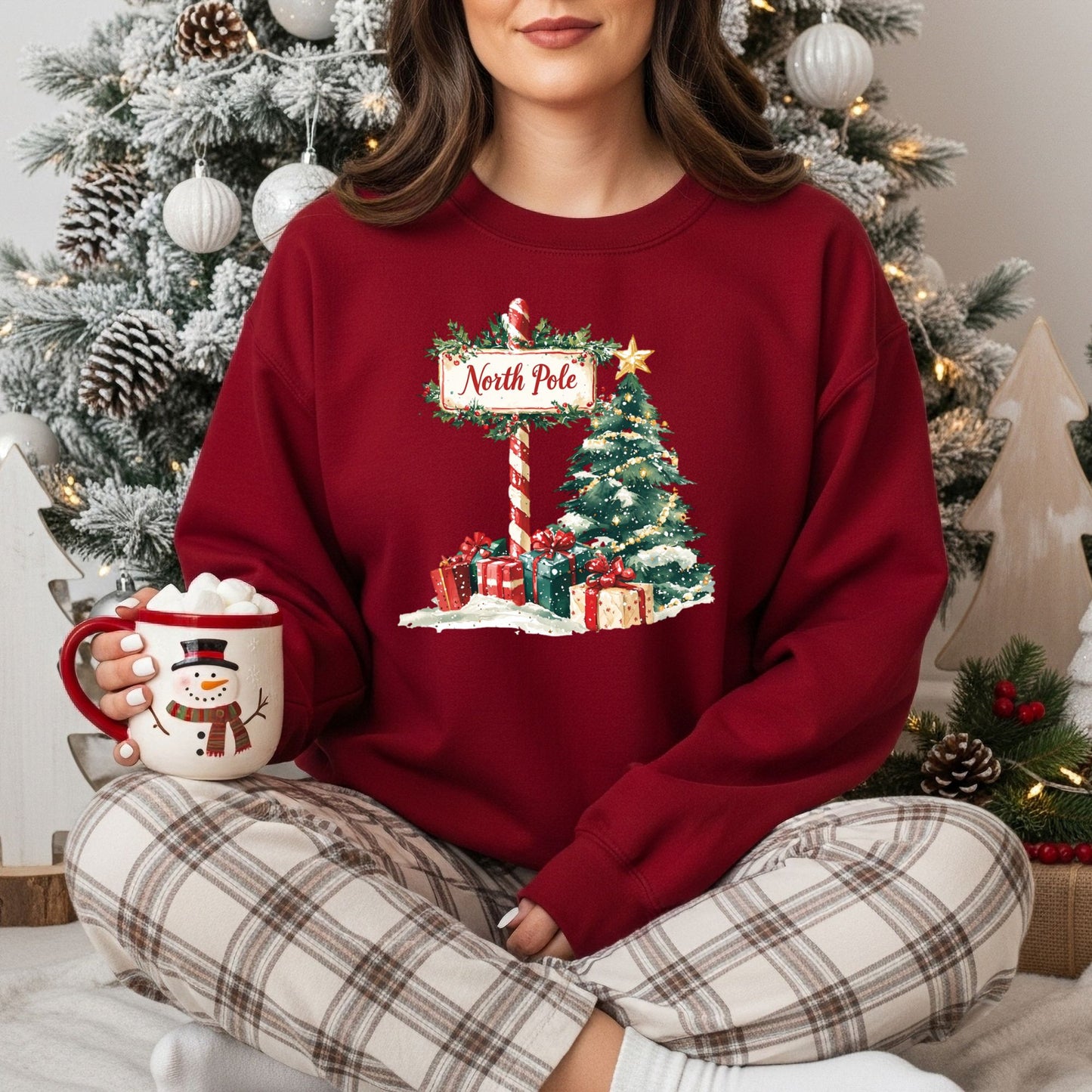 Red North Pole Christmas sweatshirt with watercolor tree, gifts, and sign, worn by a woman holding a snowman mug.