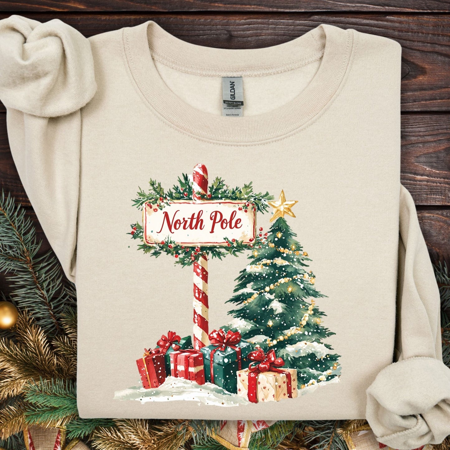Sand Gildan 18000 sweatshirt with a watercolor North Pole sign, snowy Christmas tree, and festive gifts.