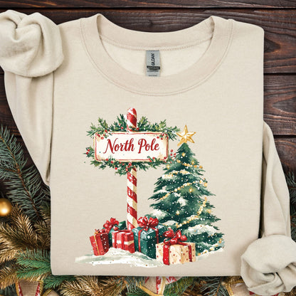 Sand Gildan 18000 sweatshirt with a watercolor North Pole sign, snowy Christmas tree, and festive gifts.