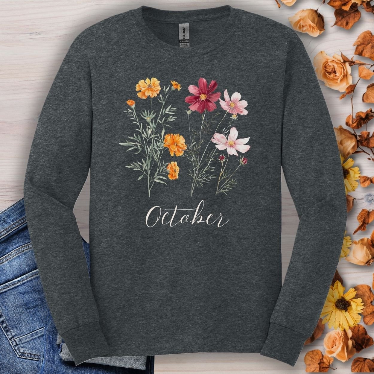 Dark Heather long sleeve tee with watercolor marigolds and cosmos, "October" text, on a wooden surface with autumn leaves.