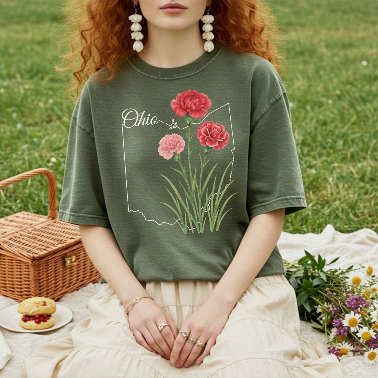 Garment-dyed olive green t-shirt with a white Ohio outline and red and pink carnations, worn by a person sitting on a picnic blanket.