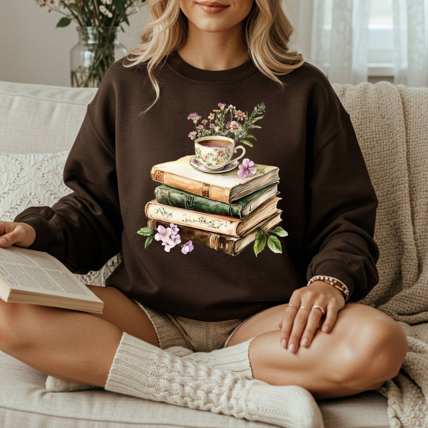 Woman in a brown crewneck sweatshirt with a vintage illustration of stacked books, a floral teacup, and greenery.