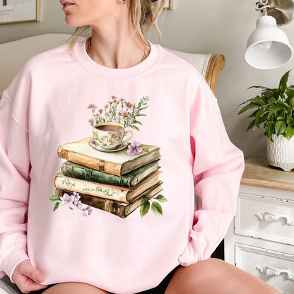 Pink crewneck sweatshirt with a vintage illustration of stacked books, a floral teacup, and greenery.