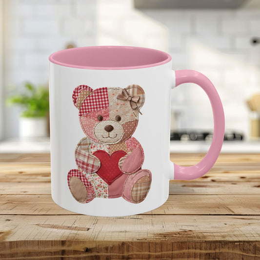 White 11oz ceramic mug with a pink interior and handle, featuring a patchwork teddy bear holding a red heart.