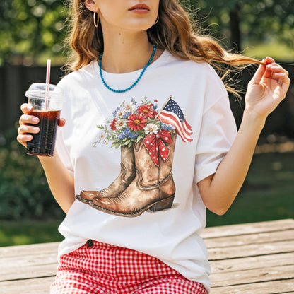 White t-shirt with watercolor cowboy boots, vibrant flowers, an American flag, and star ribbon.