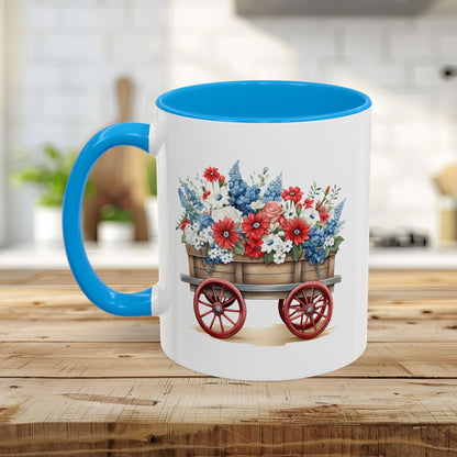 White ceramic mug with blue handle/interior, featuring a vintage wagon overflowing with red, white, and blue florals.