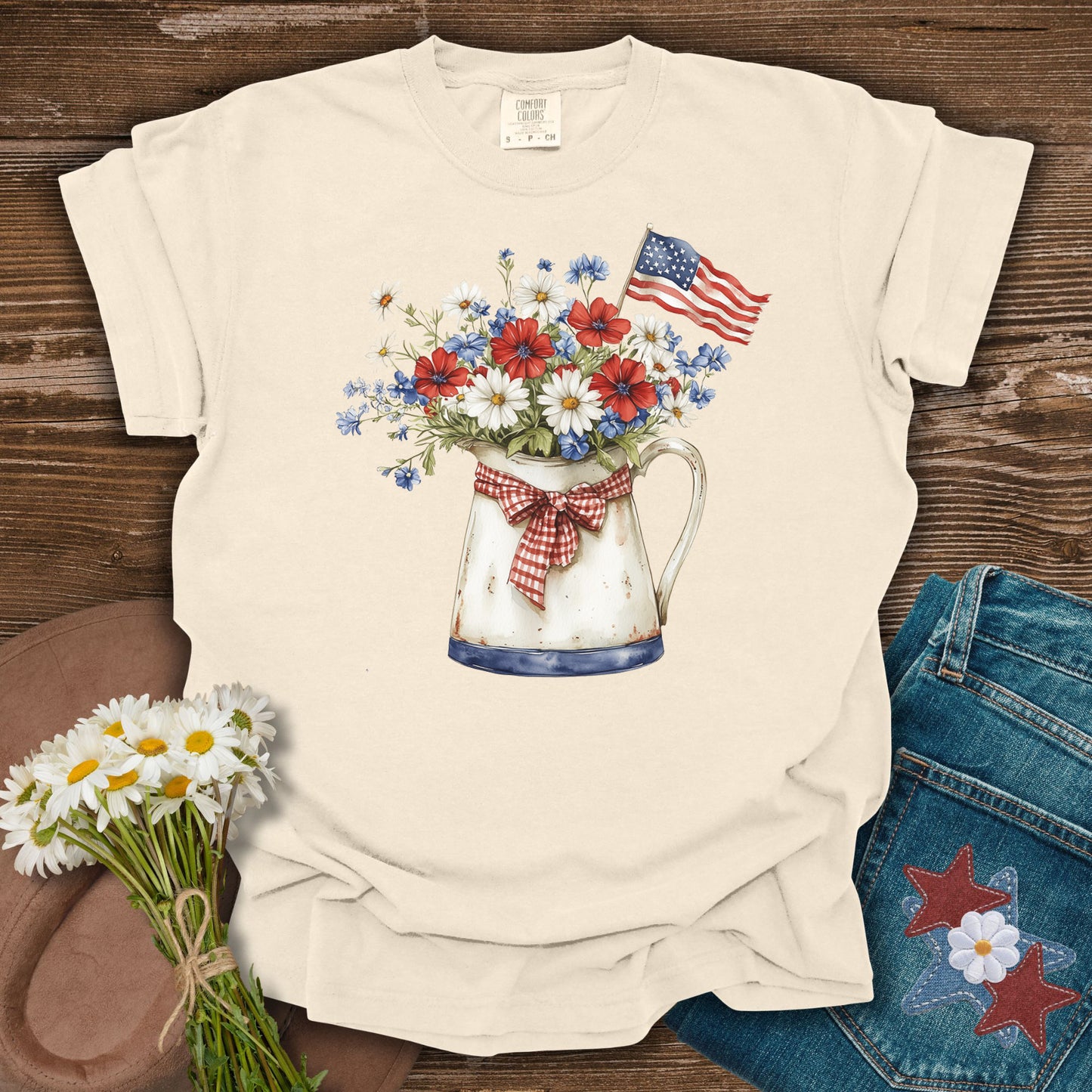 Beige t-shirt with a floral design and American flag on a wooden surface with flowers and jeans.