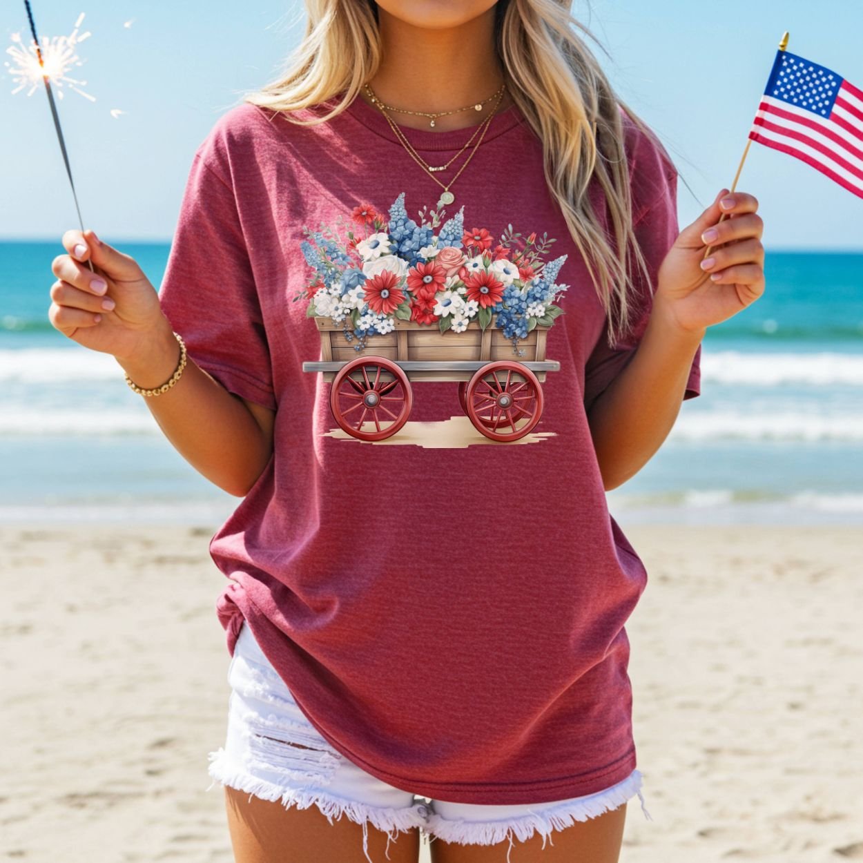 Garment-dyed red cotton t-shirt with a patriotic flower wagon design, worn by a person holding a sparkler and US flag on a beach.