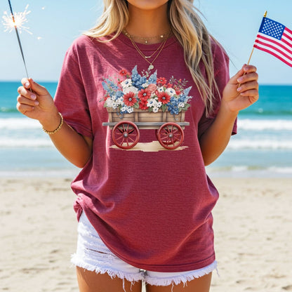 Garment-dyed red cotton t-shirt with a patriotic flower wagon design, worn by a person holding a sparkler and US flag on a beach.