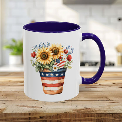 White ceramic 11oz mug with blue interior/handle, featuring a patriotic sunflower, poppy, and blue blossom bouquet in a striped pot.