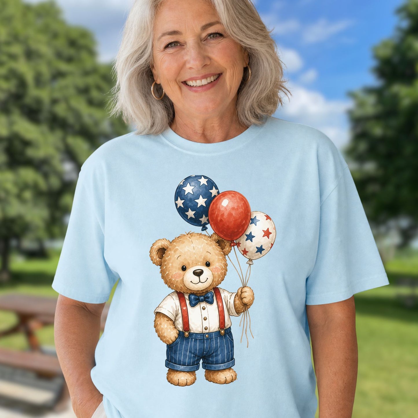 Smiling woman in a light blue tee with a vintage teddy bear holding red, white, and blue star balloons.