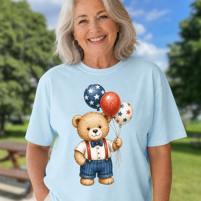 Smiling woman in a light blue tee with a vintage teddy bear holding red, white, and blue star balloons.