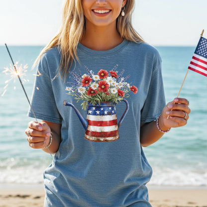 Woman in blue t-shirt with patriotic watering can overflowing with lavender, daisies, and red poppy-like flowers.